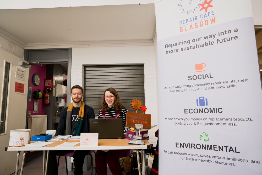 Volunteers sitting at a reception table next to a repair cafe glasgow poster
